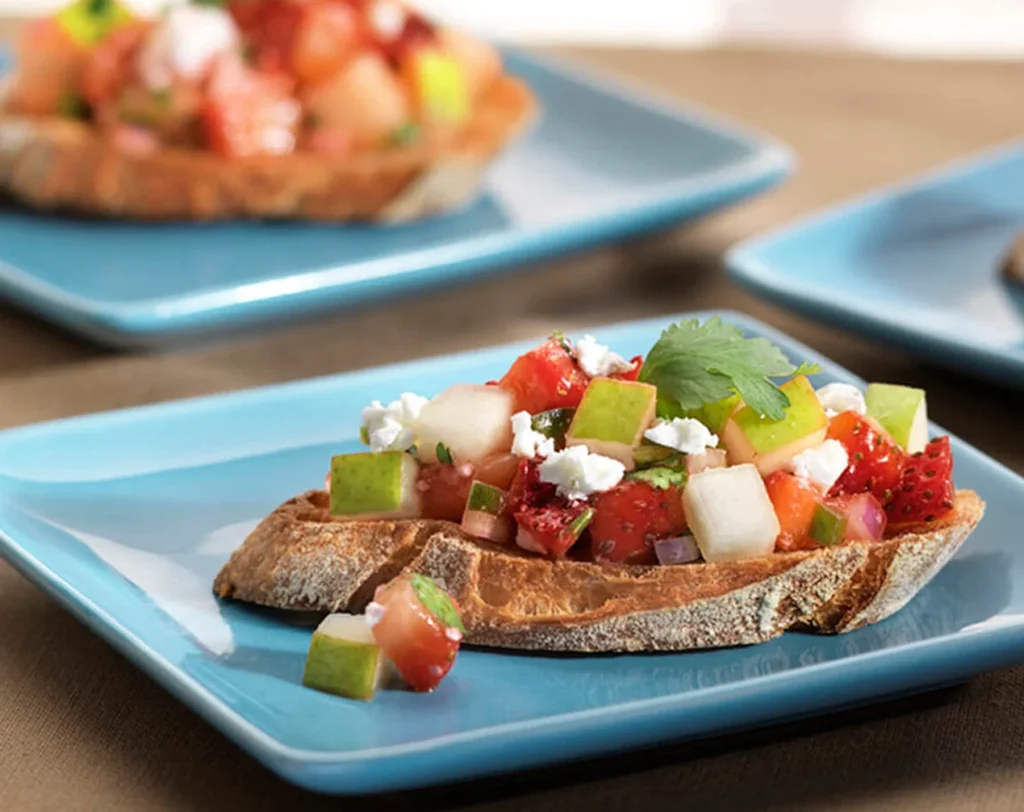 Crostini topped with pear strawberry salsa, feta cheese, cilantro, and diced fruit on toasted bread plates