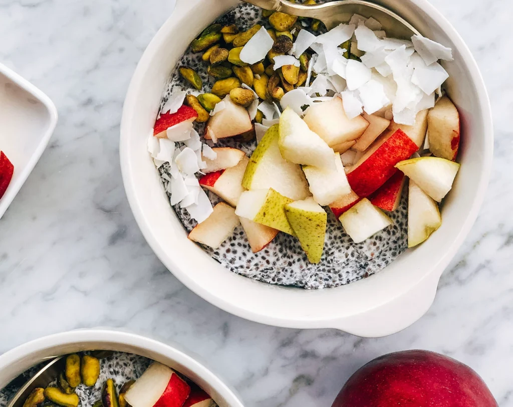 Coconut pear chia pudding topped with fresh pear, apple slices, pistachios, and coconut flakes in a white bowl