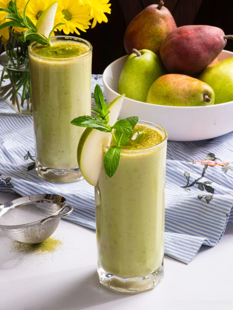 Two glasses of pear matcha smoothie garnished with pear slices and mint, with a bowl of pears and yellow flowers in the background