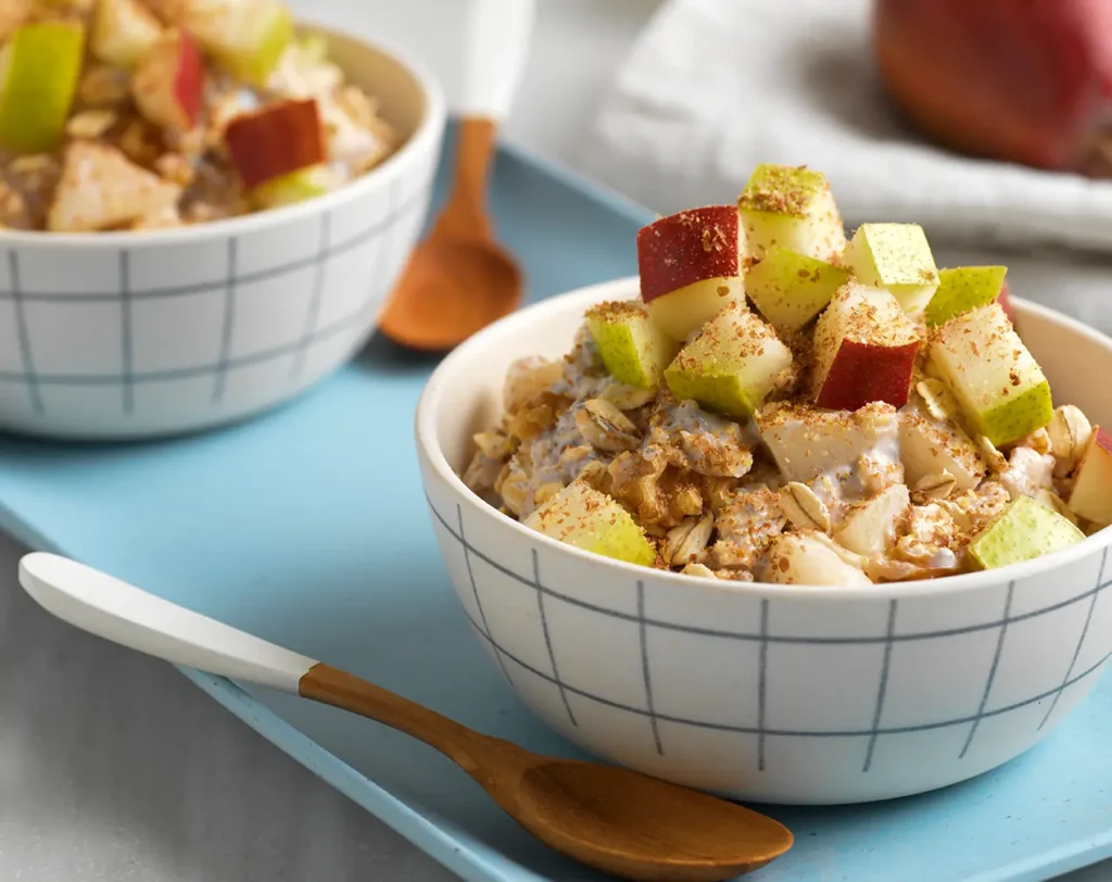 Two bowls of overnight oats topped with diced pear and red-skinned fruit, sprinkled with cinnamon, on a blue tray with wooden spoons.
