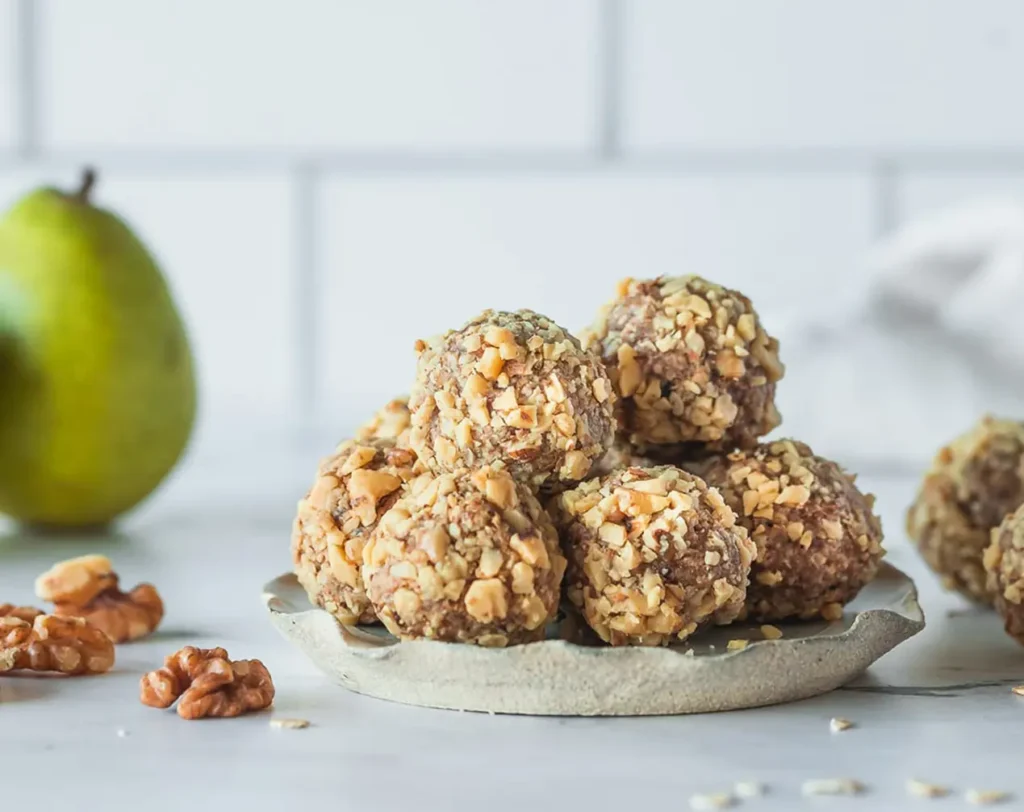 Pear, walnut, and oat energy bites coated in chopped walnuts on a small plate, with a pear and walnut pieces in the background.