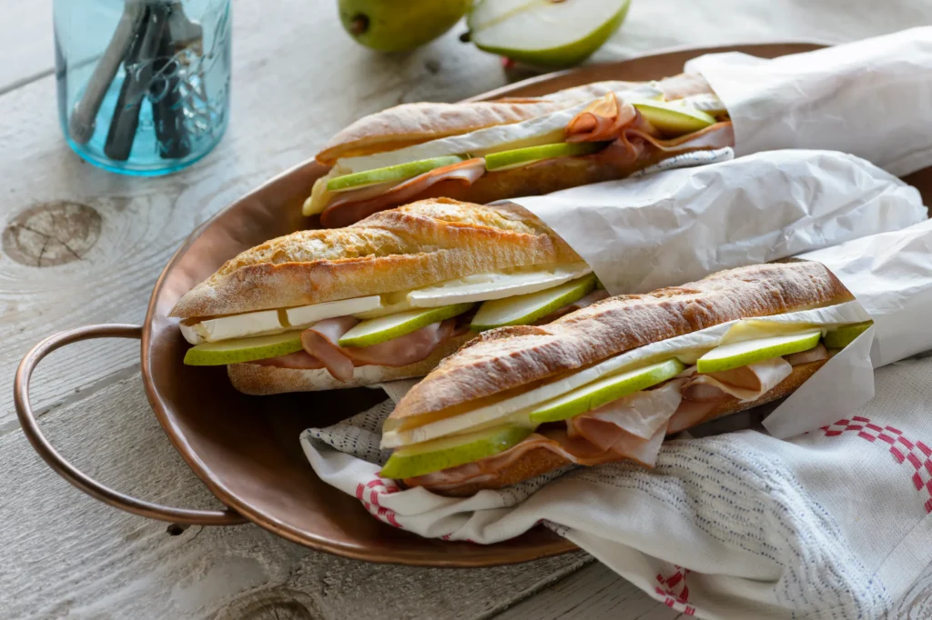 Baguette sandwiches with shaved ham, sliced pear, and brie on a copper tray.